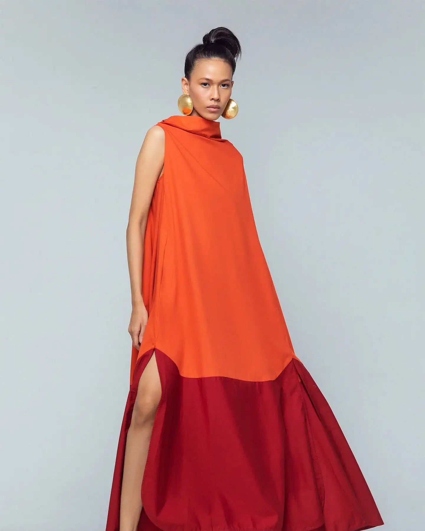 Woman in sleeveless orange and red maxi dress with side slit, gold earrings, red sandals