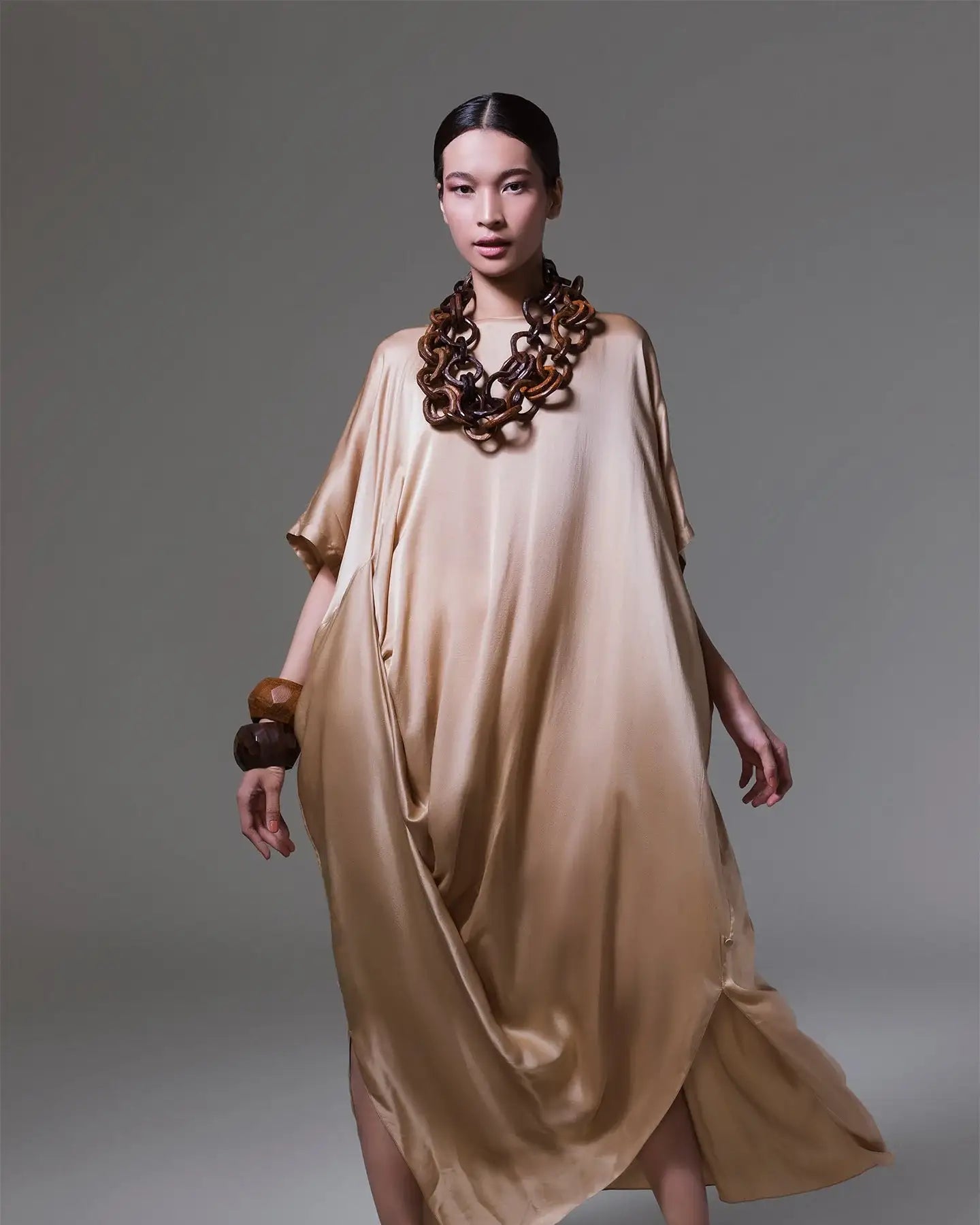 Woman in a loose gold satin dress with chunky wooden jewelry and black sandals, studio background