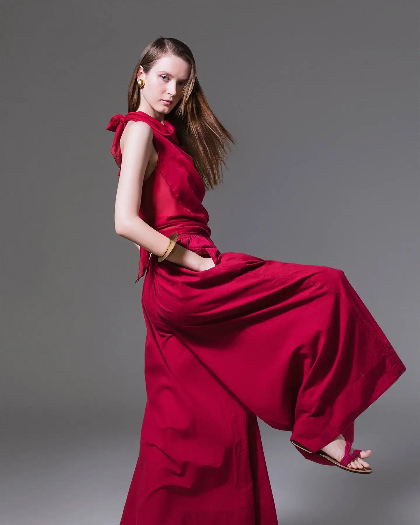 Woman in a sleeveless red wide-leg jumpsuit and sandals posing on a grey background