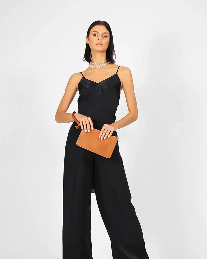 Woman in black sleeveless jumpsuit, holding tan clutch, wearing sandals and statement necklace
