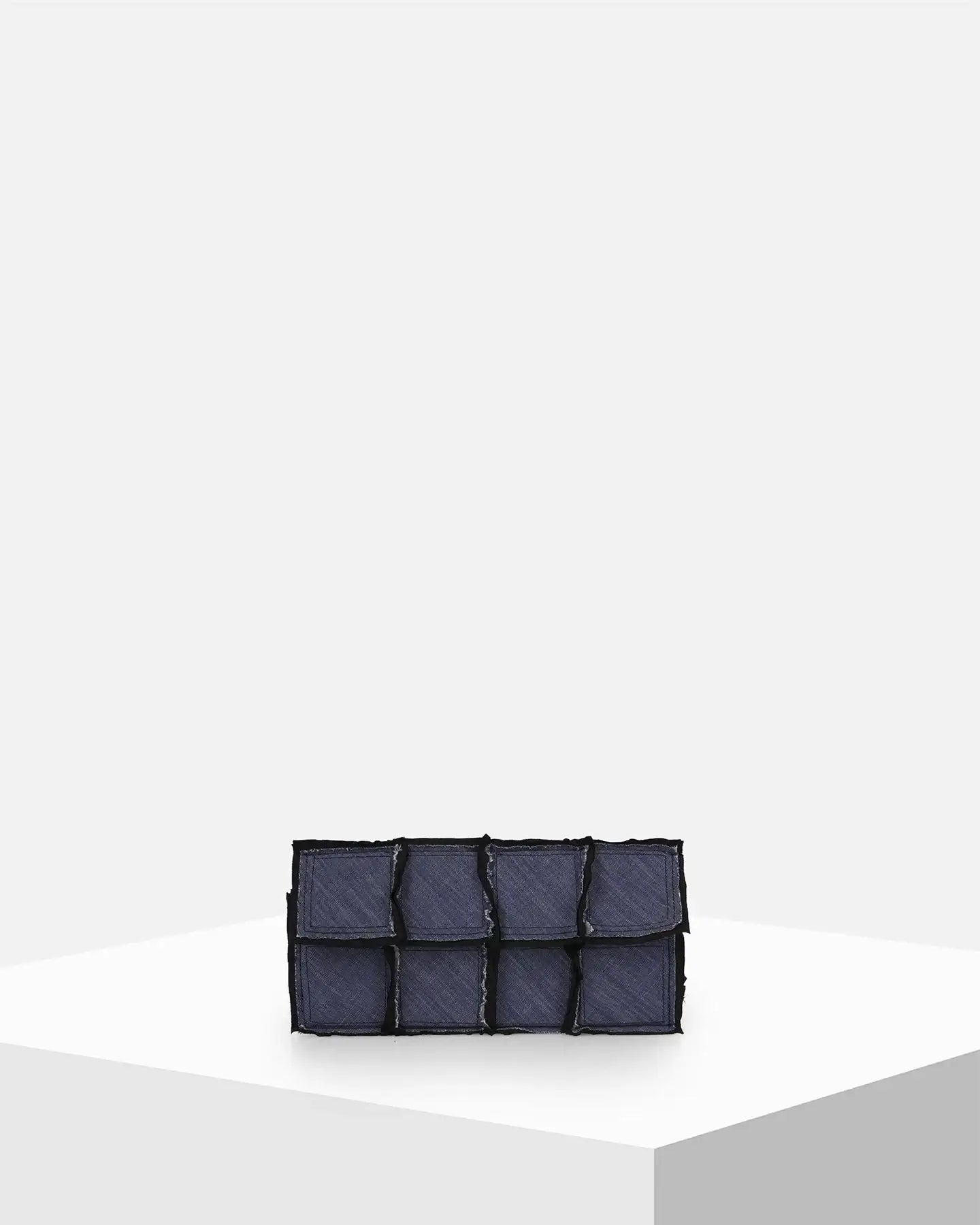 Dark denim patchwork clutch bag with textured pockets on a minimalist white pedestal