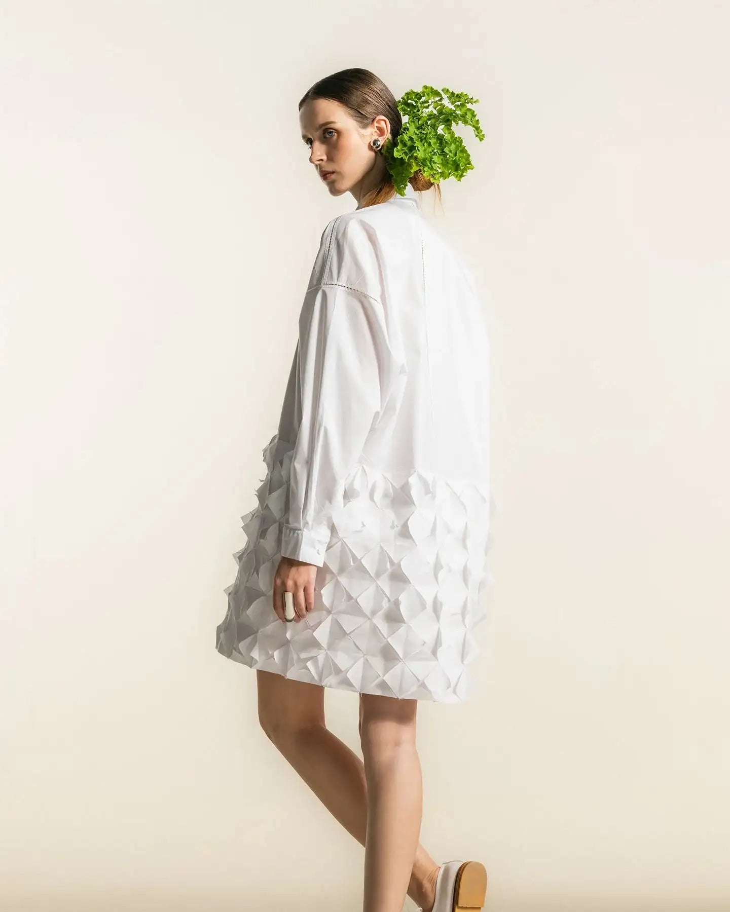 Woman in white textured geometric dress, minimal background, wearing white shoes, creative hairstyle with greenery.