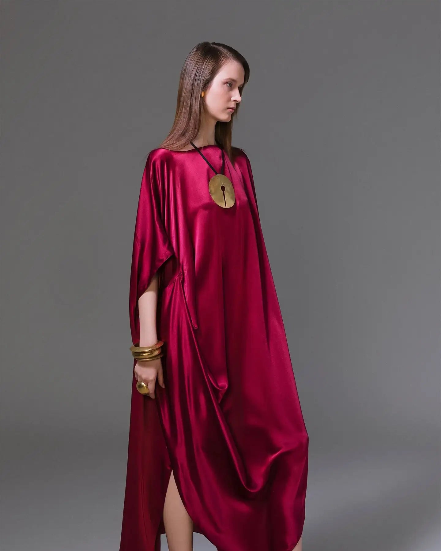 Woman in a burgundy satin kaftan dress with gold jewelry and matching sandals, studio shot