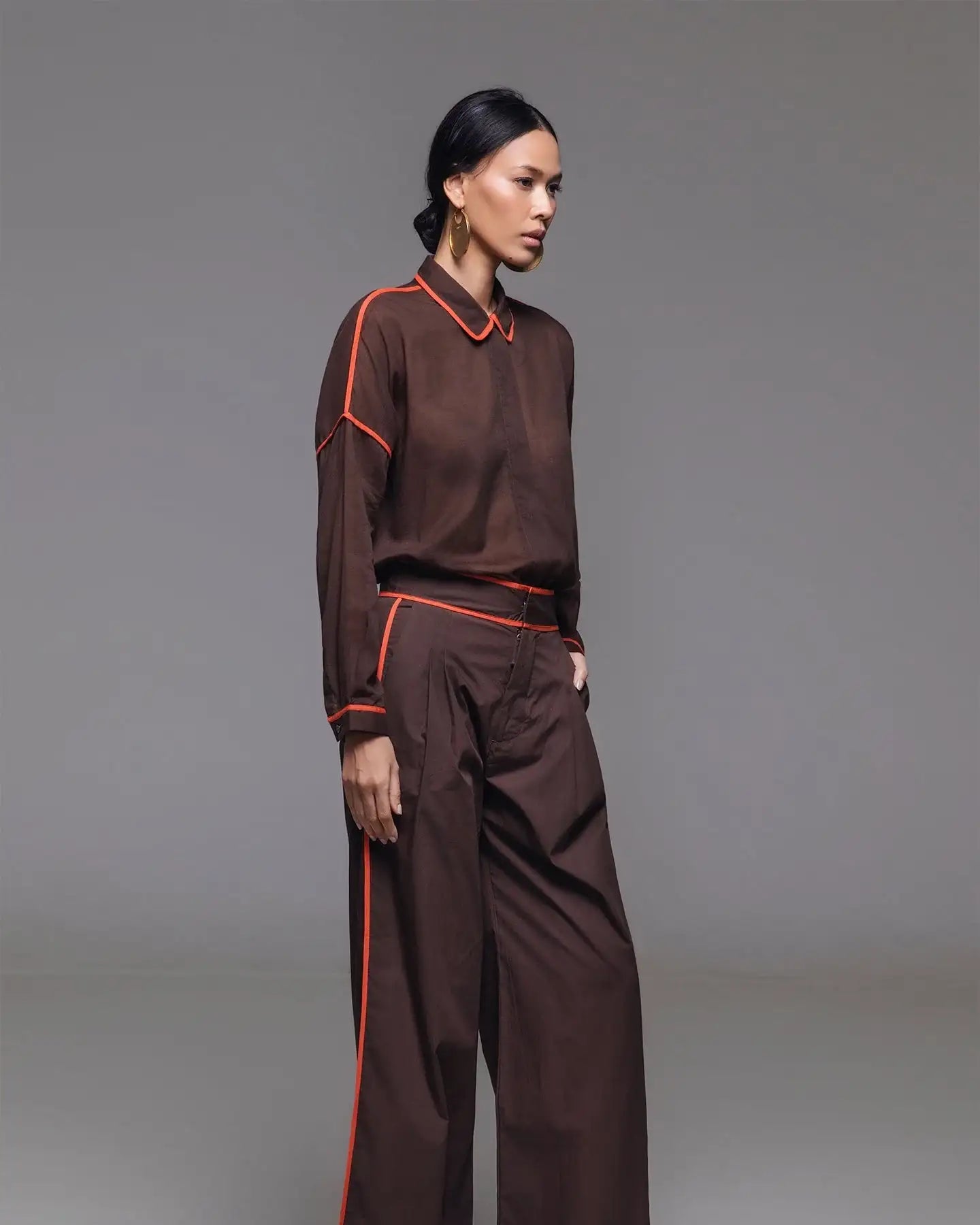 Woman in brown wide-leg pants and matching top with red piping, gold flats, and gold earrings