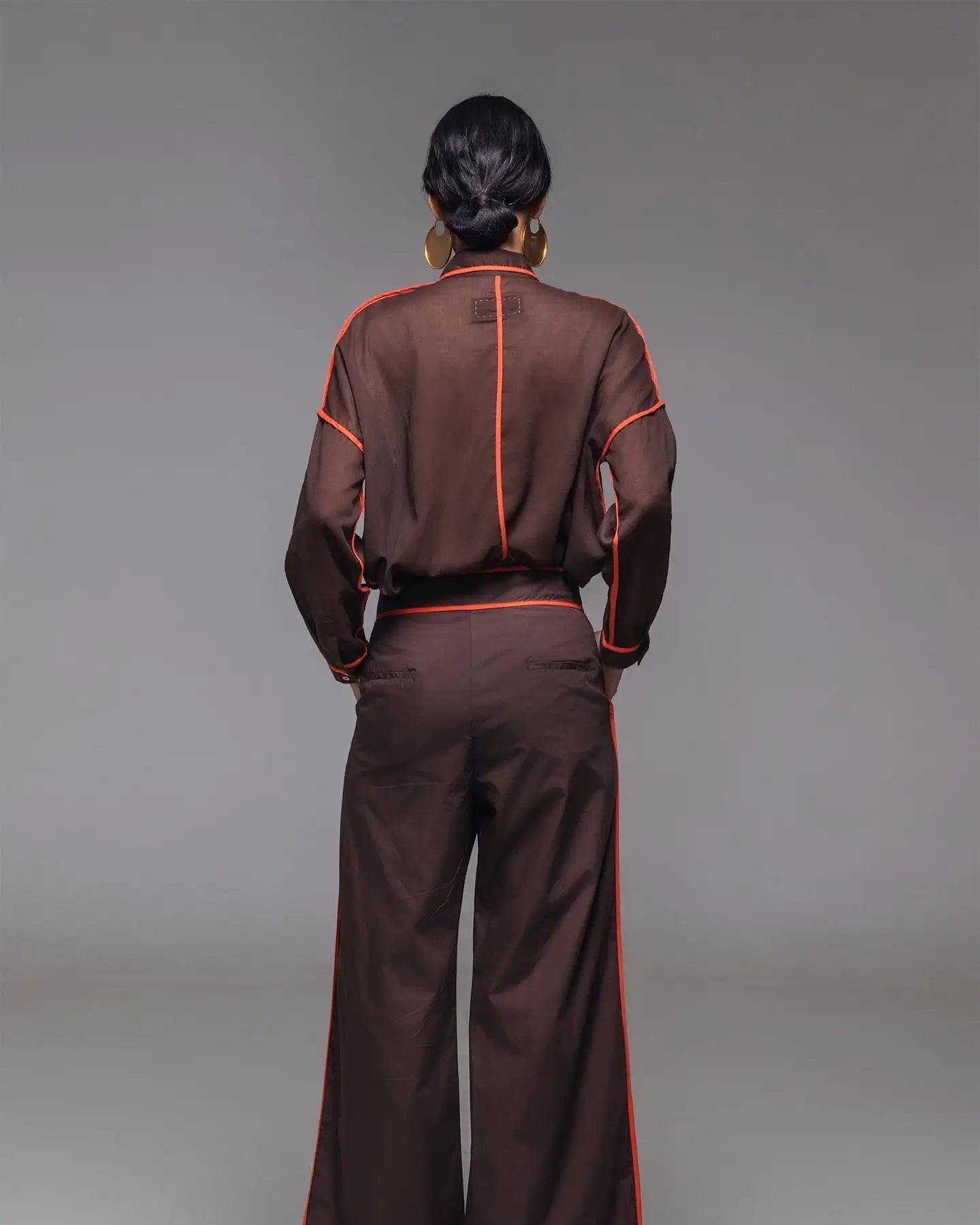 Woman in brown sheer blouse and wide-leg trousers with red piping, gold earrings, gold shoes