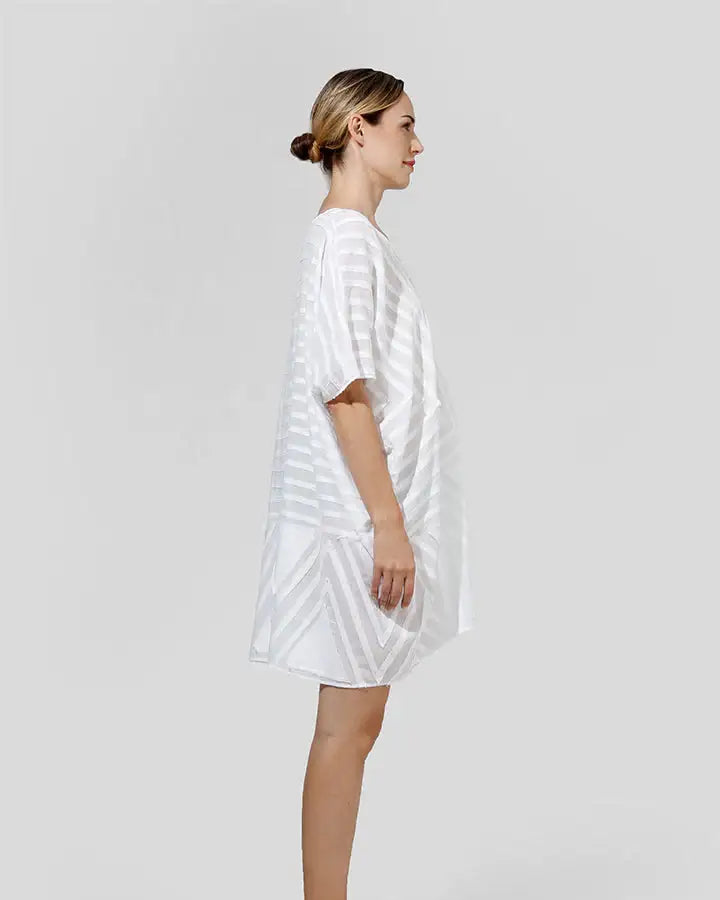 Woman in loose white geometric short dress, standing sideways on plain background