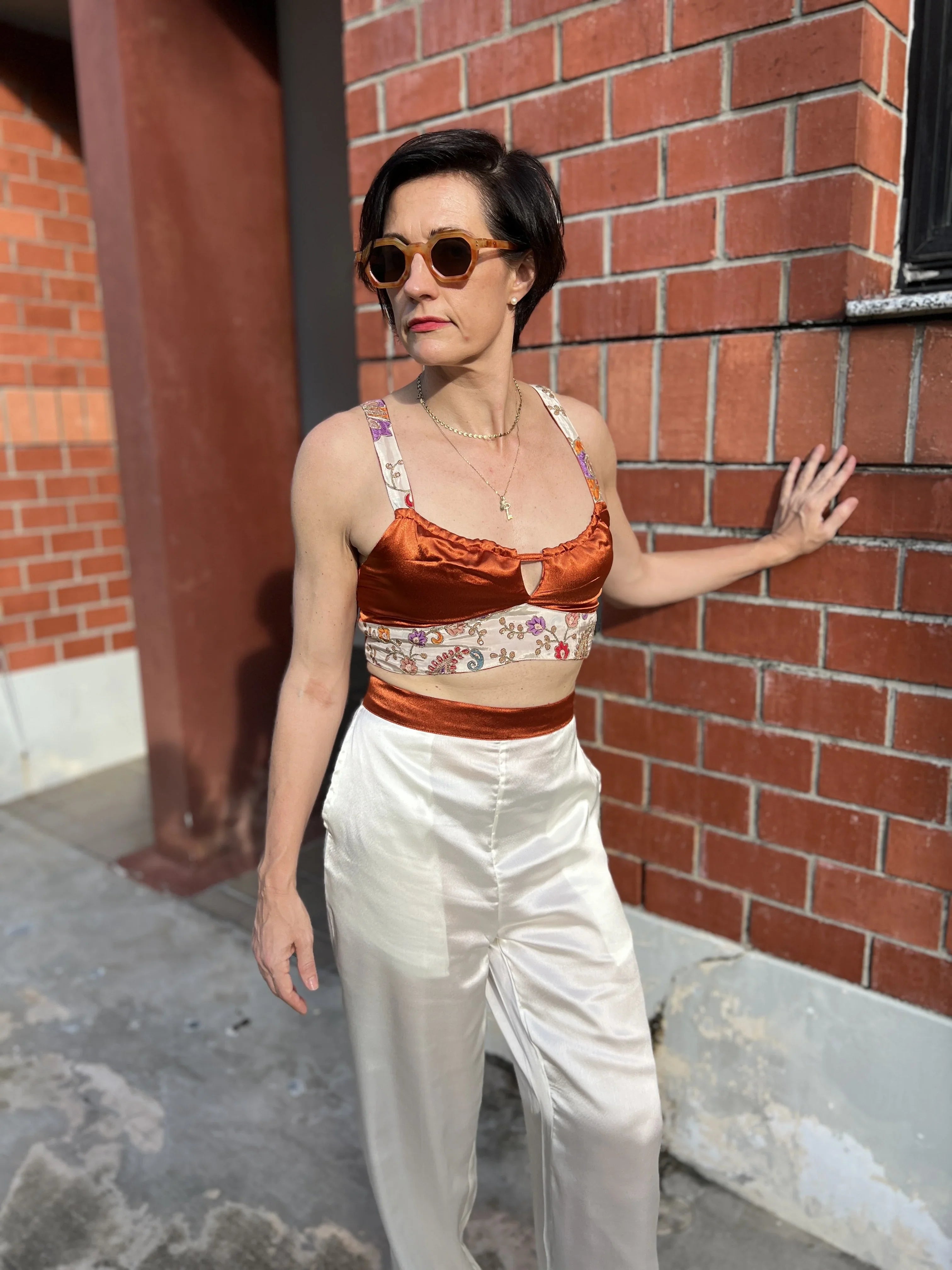 Woman in sunglasses wearing a satin crop top and white silk pants by a brick wall