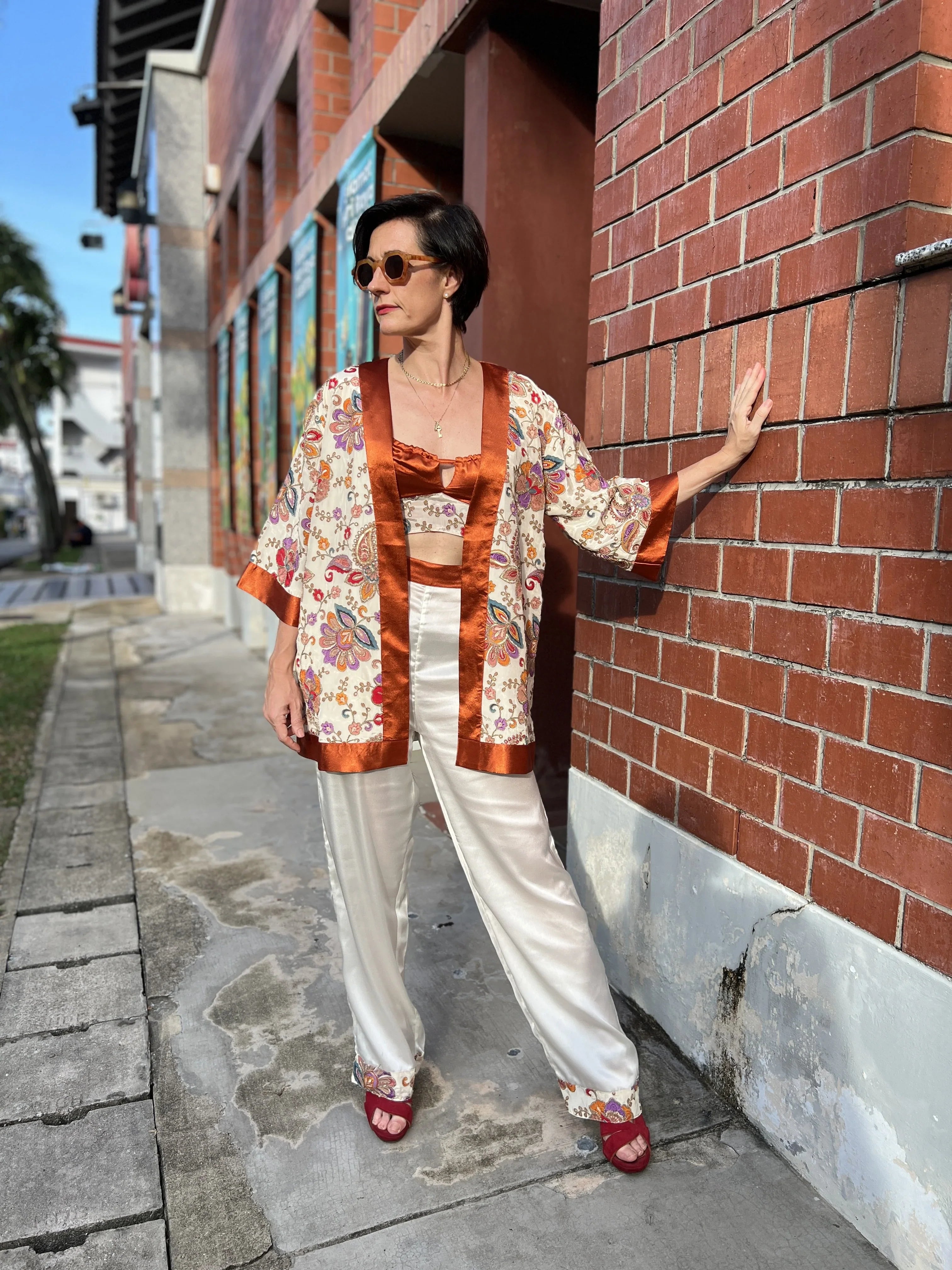 Woman in floral satin kimono top and matching pants, posing outdoors by brick wall.