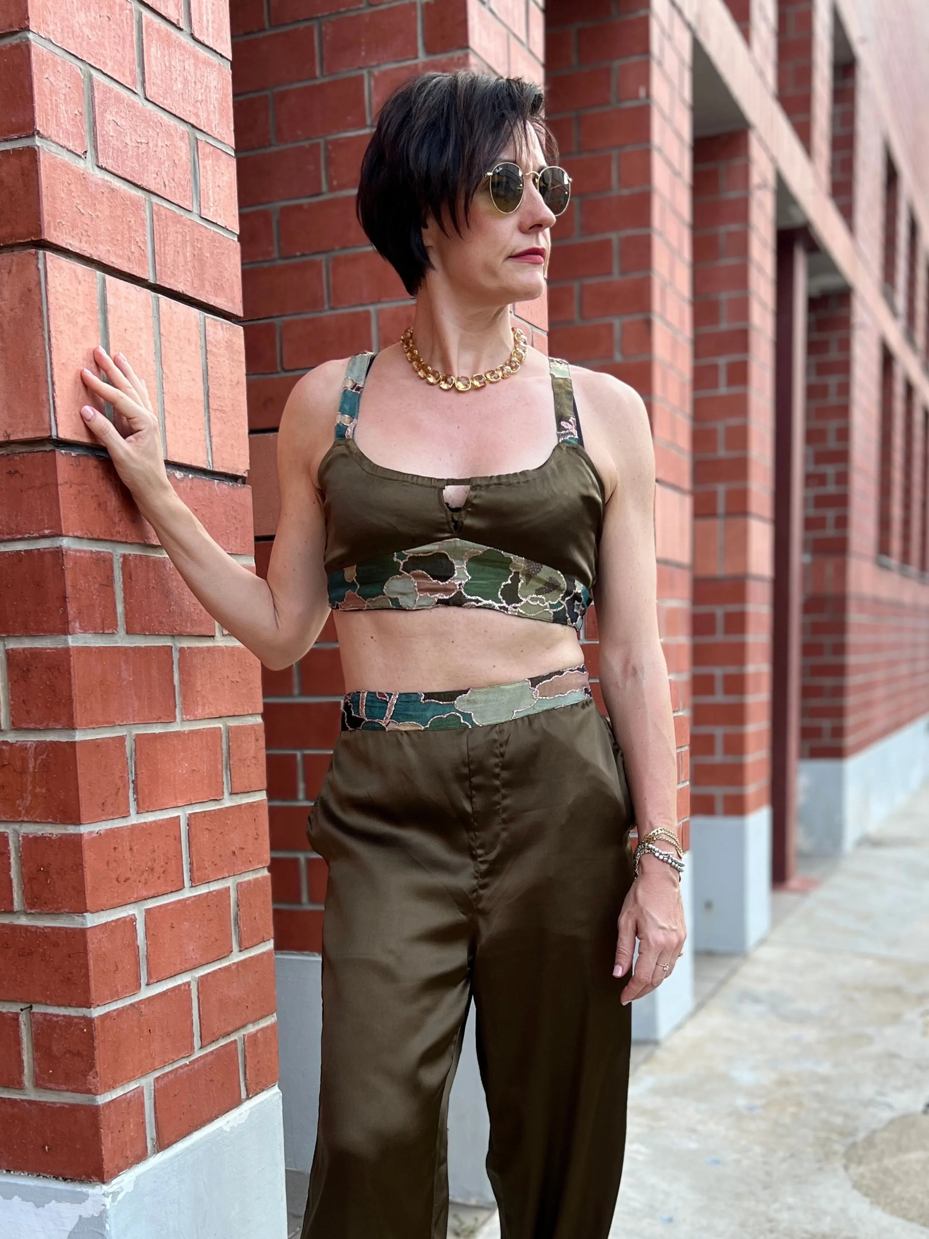 Woman in green crop top and pants with camo accents, standing by brick wall, wearing sunglasses and gold jewelry