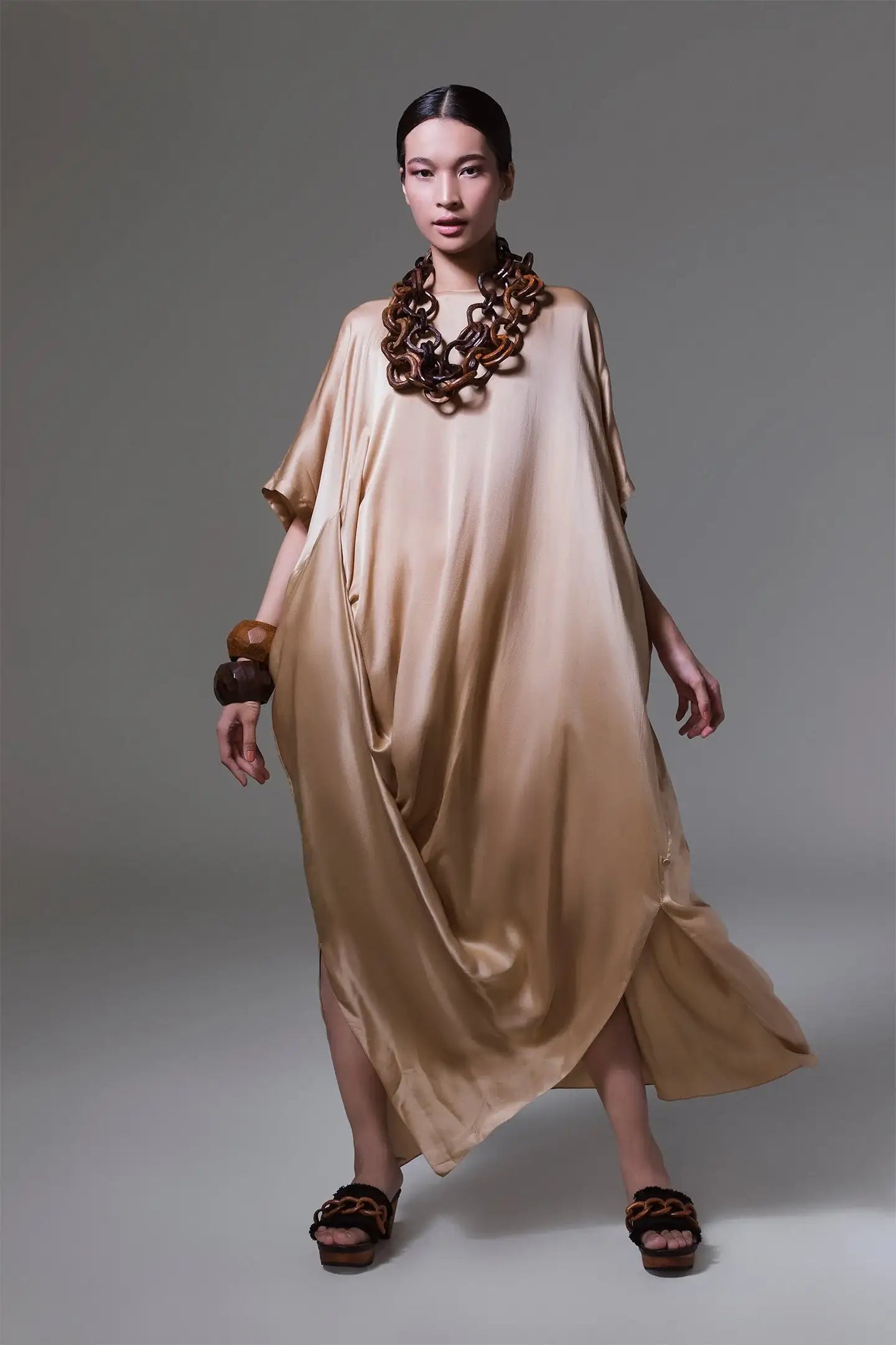 Woman in a loose gold satin dress with chunky wooden jewelry and black sandals, studio background