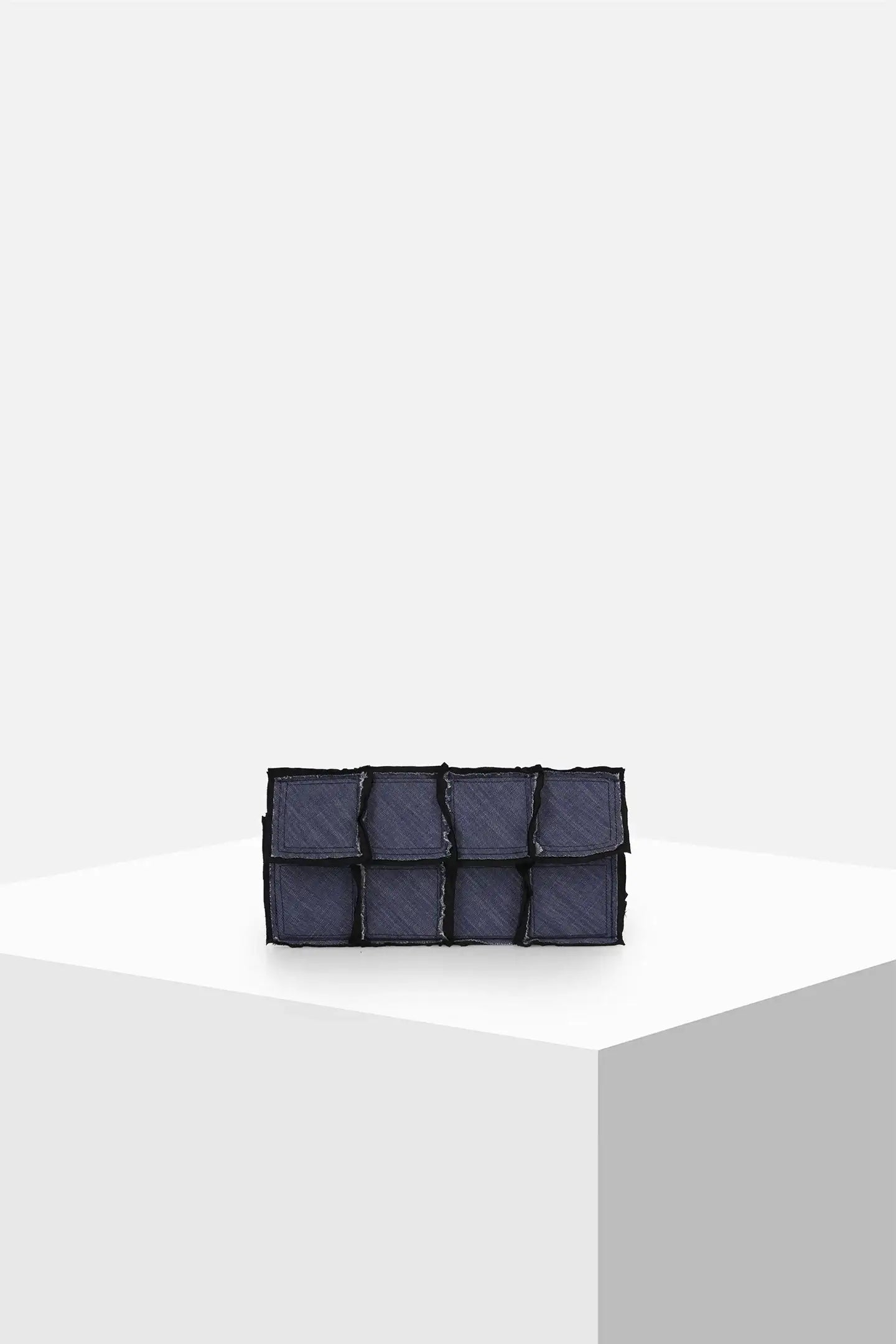 Dark denim patchwork clutch bag with textured pockets on a minimalist white pedestal