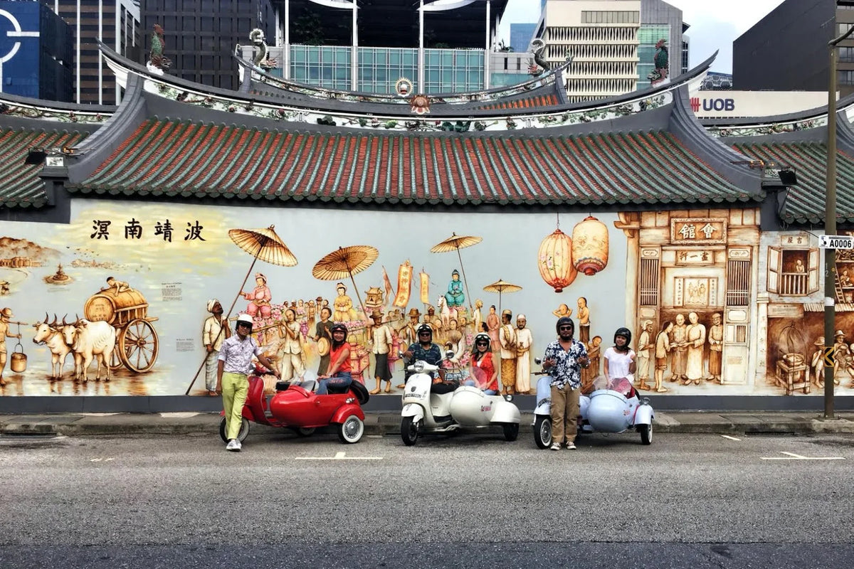 A street scene in front of a large illustrated mural on a traditional tiled roof building. Three people stand with small motorized sidecars on the road, while a red carriage and a vintage scooter are depicted in the mural behind them.